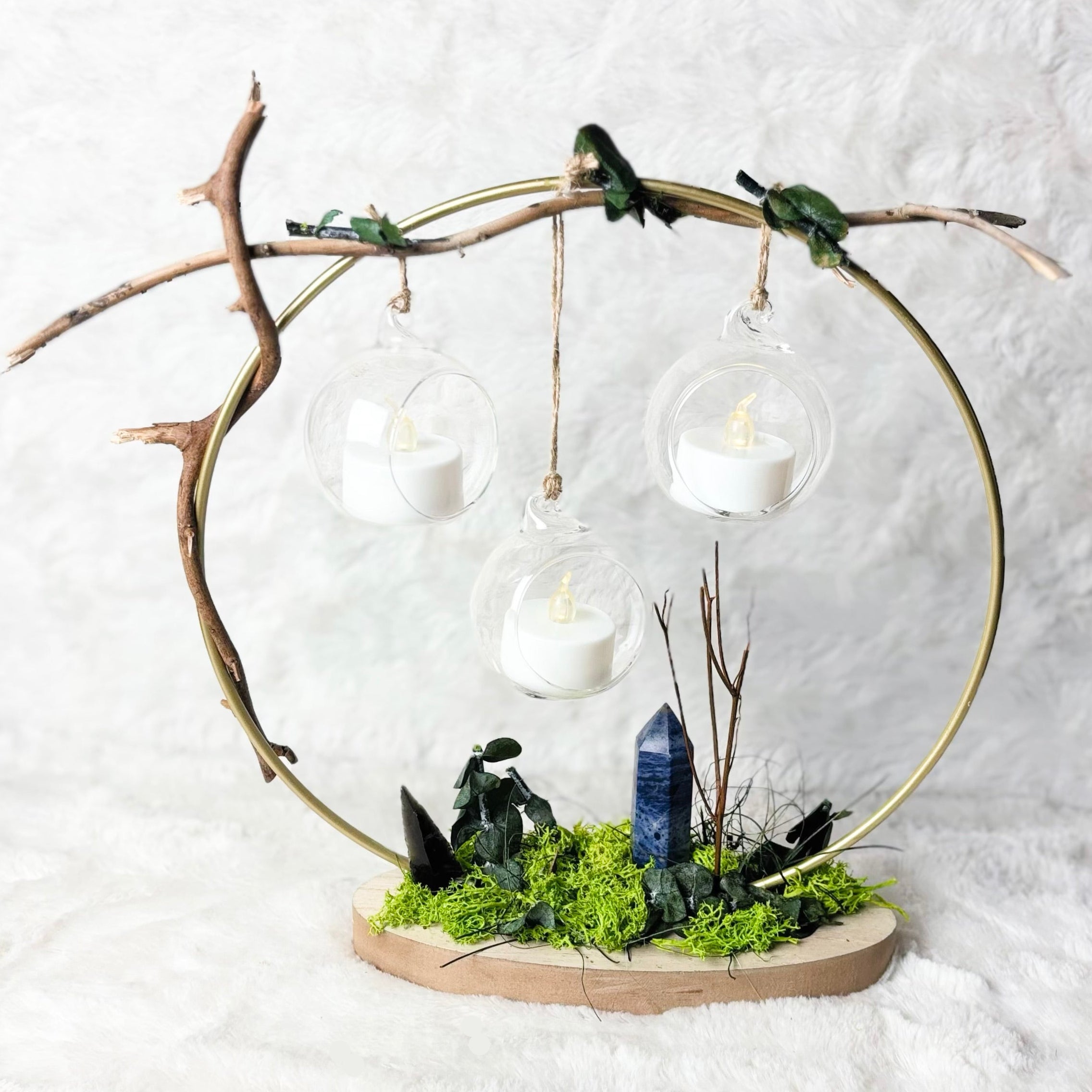 A decorative fairy garden centerpiece featuring a circular gold metal frame, small green birds perched on the branches, three hanging glass teardrop lanterns with white candles, mossy green ground, and a blue crystal tower on a natural wood base.