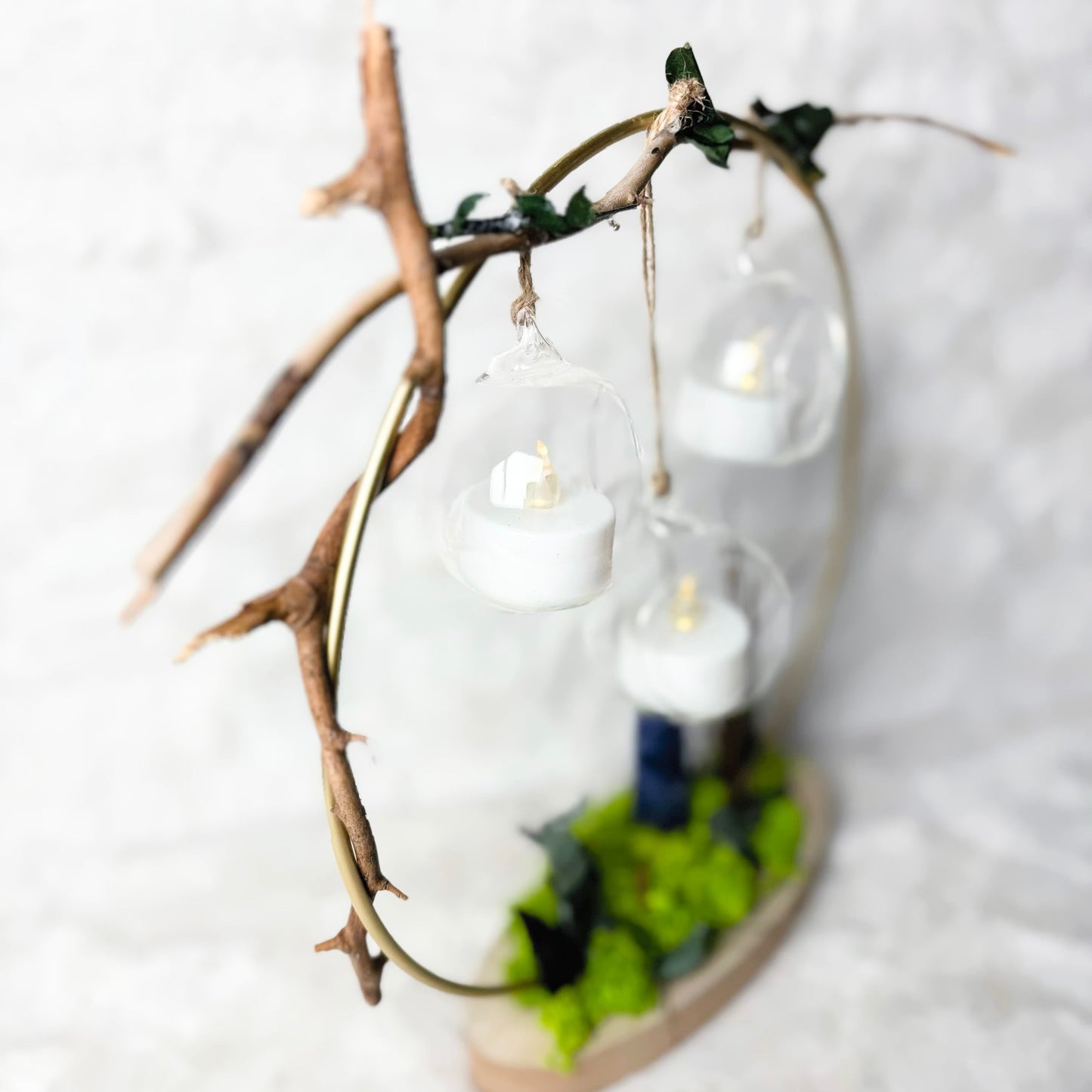 A decorative hanging light fixture made from a natural twig frame with three clear glass dome globes containing white tea light candles, set on a small base with green leaves and a dark blue accent, photographed against a soft white textured background.