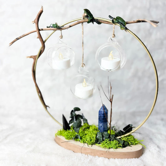 A decorative tabletop arrangement featuring a circular gold metal frame suspended from a natural branch, with three clear glass hanging lanterns containing white tea light candles. The base is covered in bright green moss and small dark stones, with a blue crystal tower in the center, set on a light wood slab against a soft white textured backdrop.