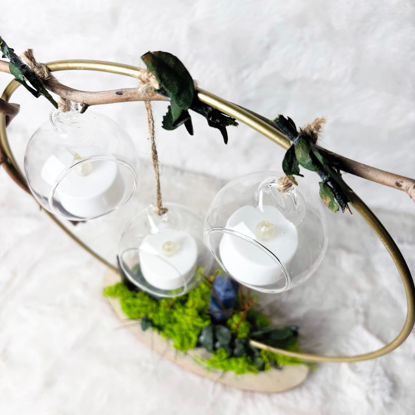 A decorative hanging candle holder made from a natural branch, featuring three clear glass orb lanterns with white tea light candles inside, accented with green moss and small dark green leaves, set against a white textured background.
