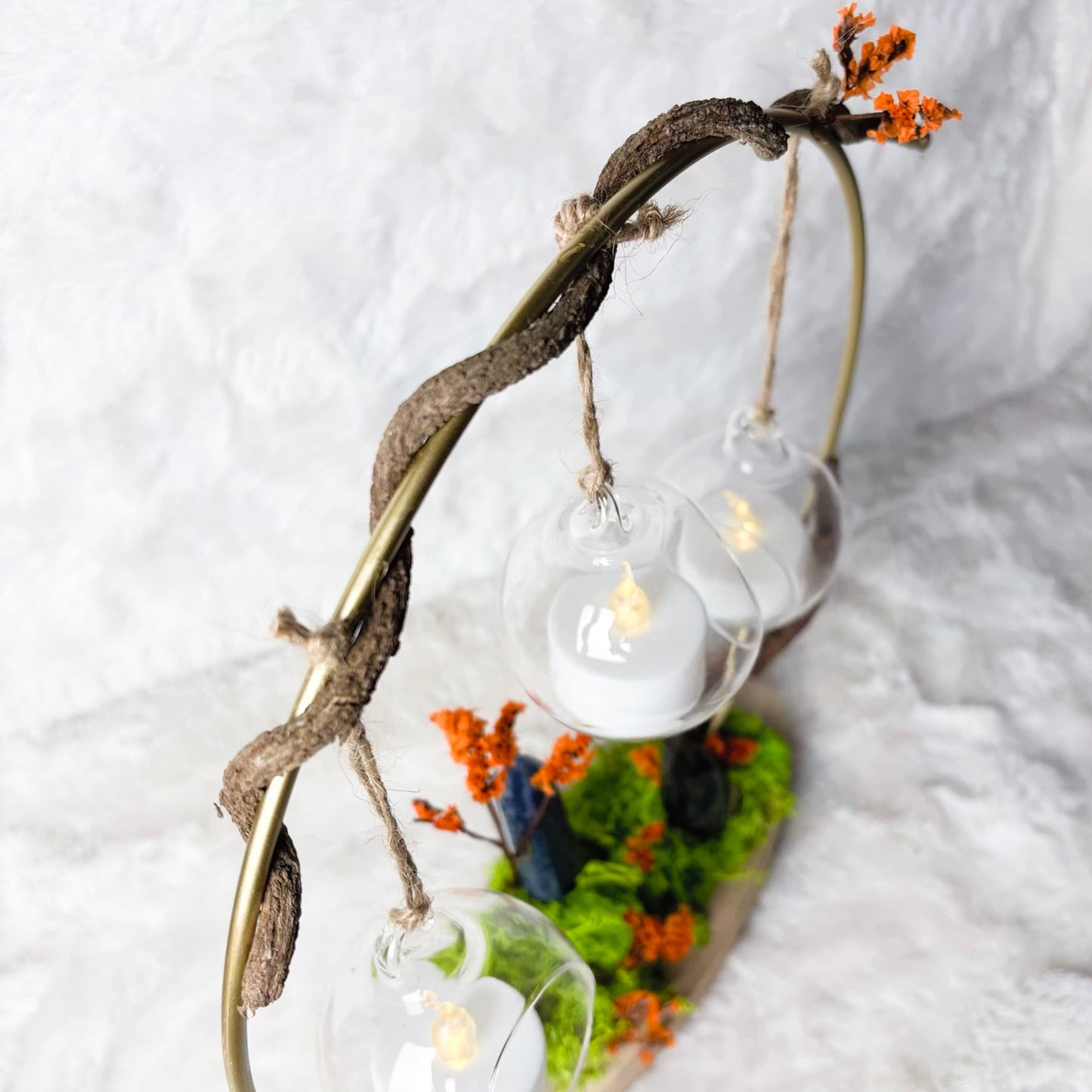 A decorative hanging light fixture with three clear glass globe bulbs suspended from a twisted dark branch frame, set against a soft white textured backdrop.