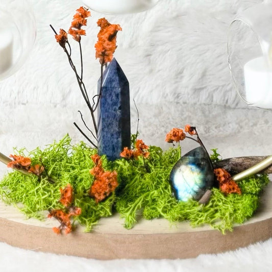 A decorative hanging glass terrarium with a circular branch frame, three clear glass cups holding white candles, mossy green ground, small orange dried flowers, and a small blue bird figurine, displayed on a white textured fabric background.
