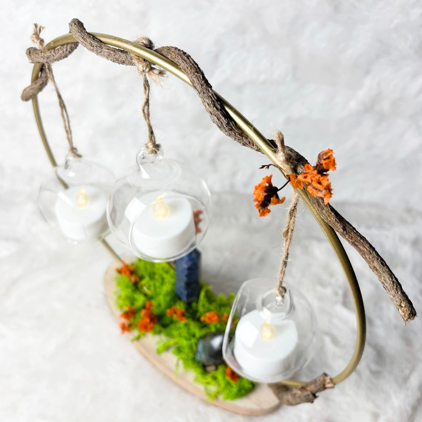 A rustic decorative hanging arrangement made from a twisted branch frame, holding three clear glass cups with white tea light candles, set on a small base with green moss and orange flowers against a soft white textured background.