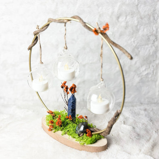 A decorative hanging lantern made from a natural driftwood circle frame, suspended over a small mossy base with tiny orange flowers, a miniature blue figure, and three clear glass globes containing white tea light candles, set against a soft white textured backdrop.