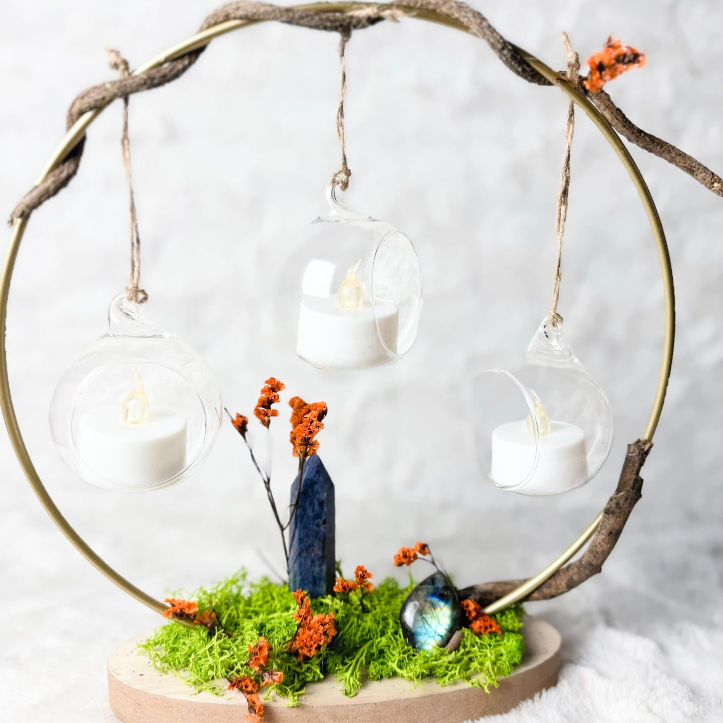 A decorative hanging candle arrangement with three clear glass orb lanterns containing white tea light candles, suspended from a twisted natural wood branch in a circular frame. The base features green moss, small orange dried flowers, a dark blue crystal, and an iridescent stone on a round wooden platform, set against a soft white textured background.