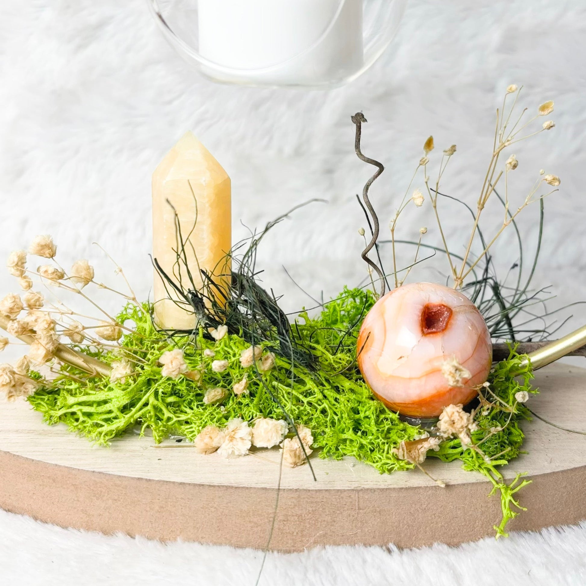 A decorative tabletop arrangement on a round wooden board, featuring green moss, dried cream flowers, a pale yellow candle, a peach-colored decorative object, and gold-toned curved rods, set against a white textured backdrop with hanging glass candle holders above.