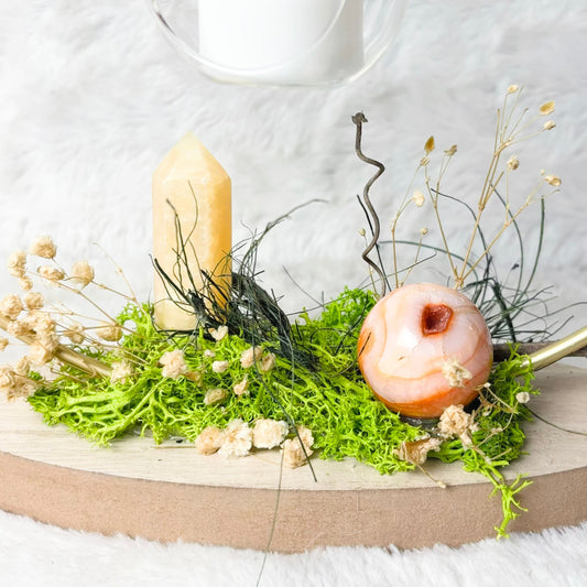 A decorative tabletop arrangement on a round wooden board, featuring green moss, dried cream flowers, a pale yellow candle, a peach-colored decorative object, and gold-toned curved rods, set against a white textured backdrop with hanging glass candle holders above.