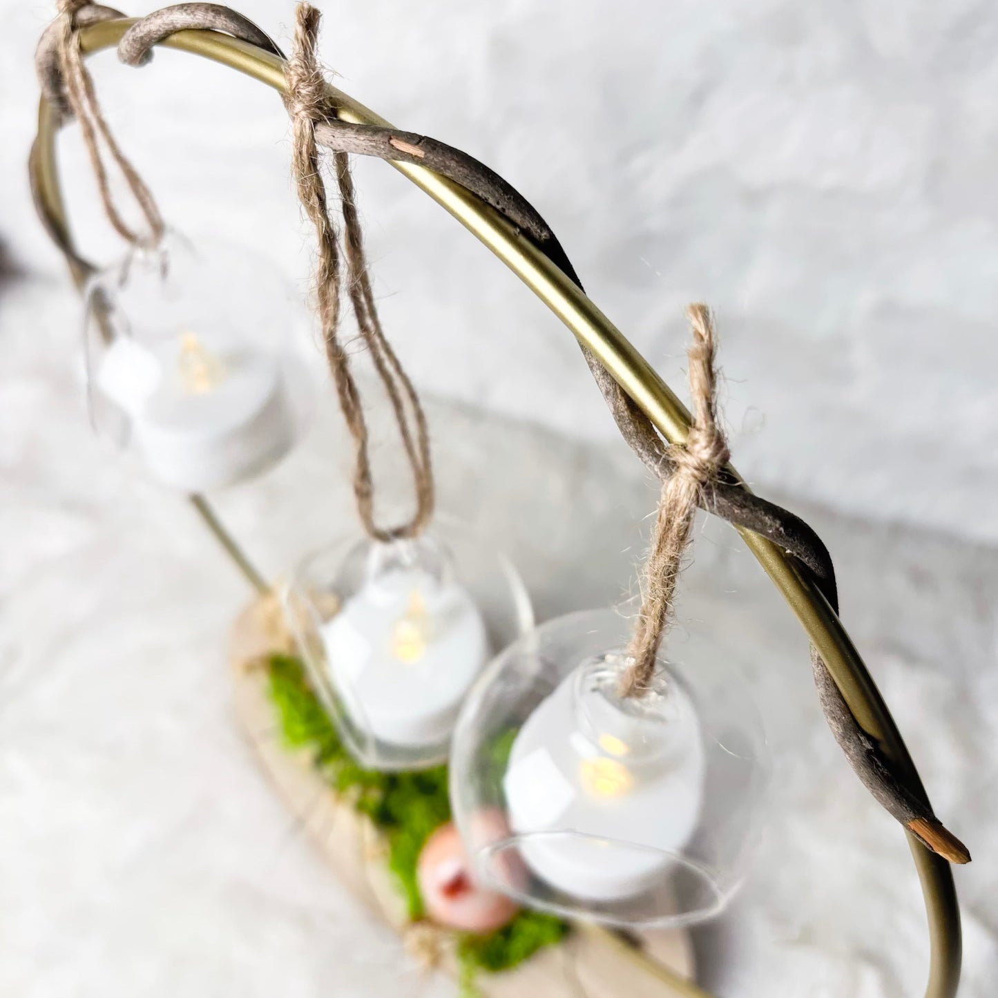 A rustic metal hanging candle holder with a curved branch-like frame, twine ties, and three clear glass cups holding white tea light candles, displayed on a light textured surface.