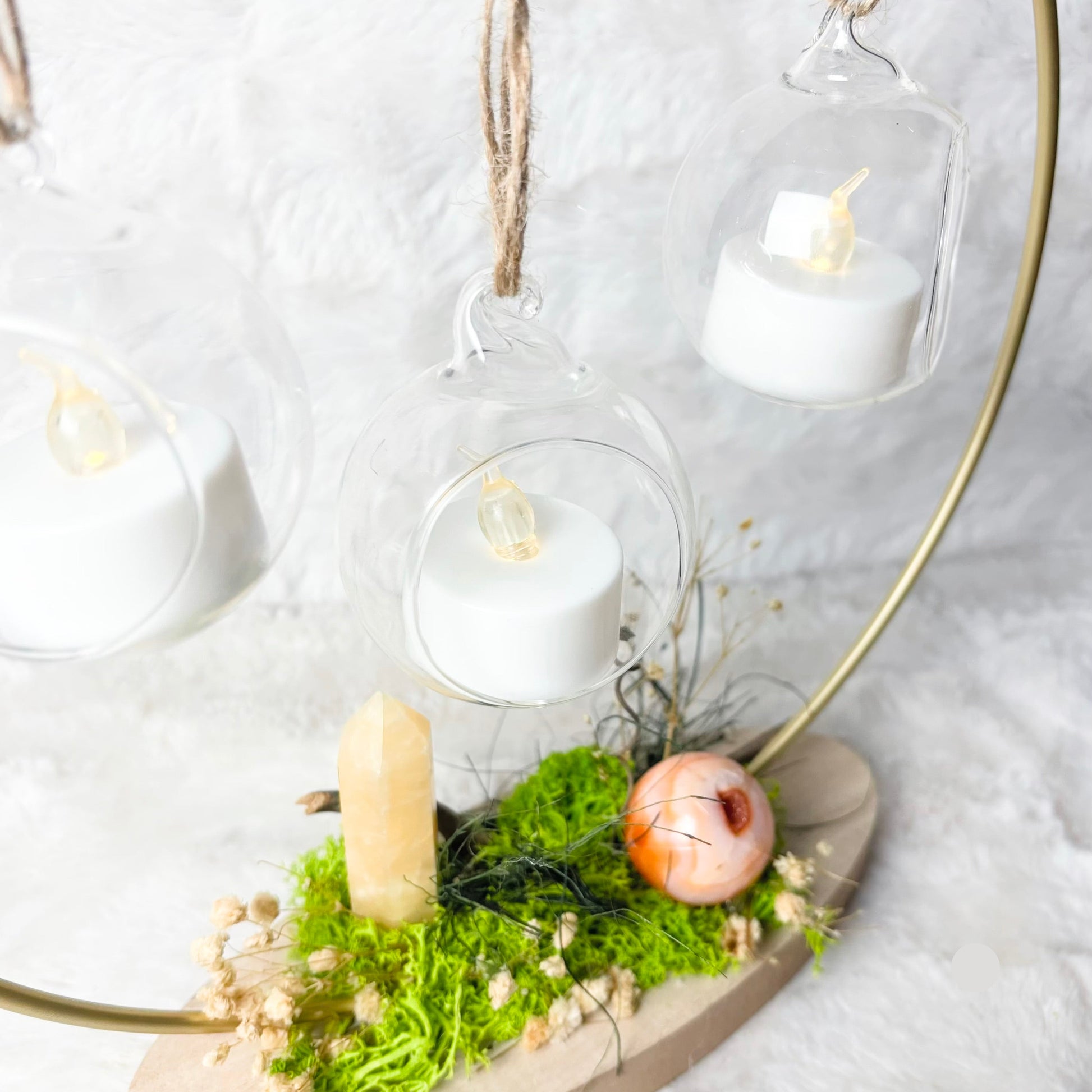 A decorative hanging candle arrangement with clear glass bell-shaped holders containing white tea light candles, suspended from a gold-toned circular frame over a wooden base with green moss, a crystal point, and dried botanicals.