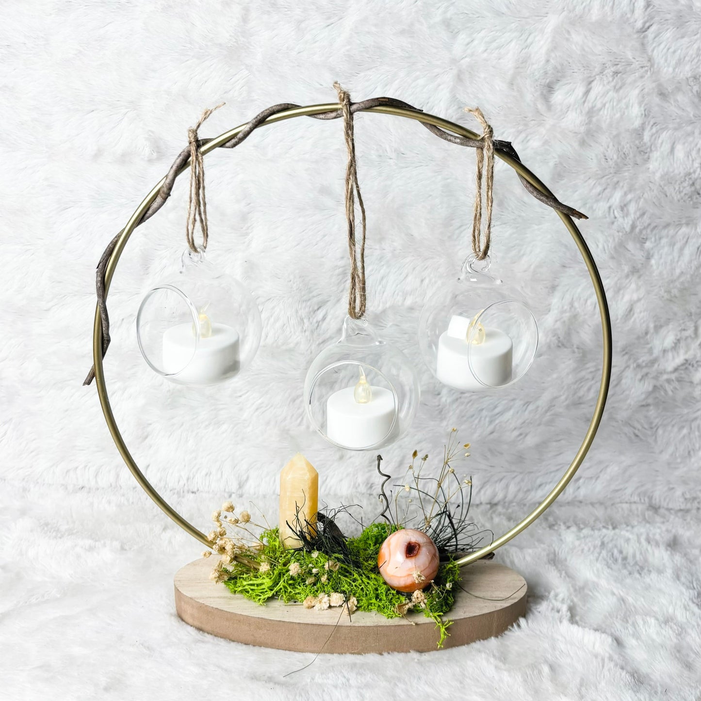 A decorative metal candle holder with a circular frame, hanging glass cups, three white tea light candles, moss, dried grass, a small candle, and a pink decorative object on a round base, set against a white textured backdrop.