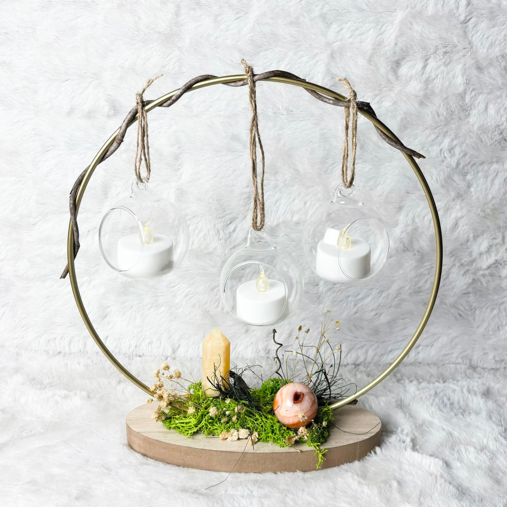 A decorative metal candle holder with a circular frame, hanging glass cups, three white tea light candles, moss, dried grass, a small candle, and a pink decorative object on a round base, set against a white textured backdrop.