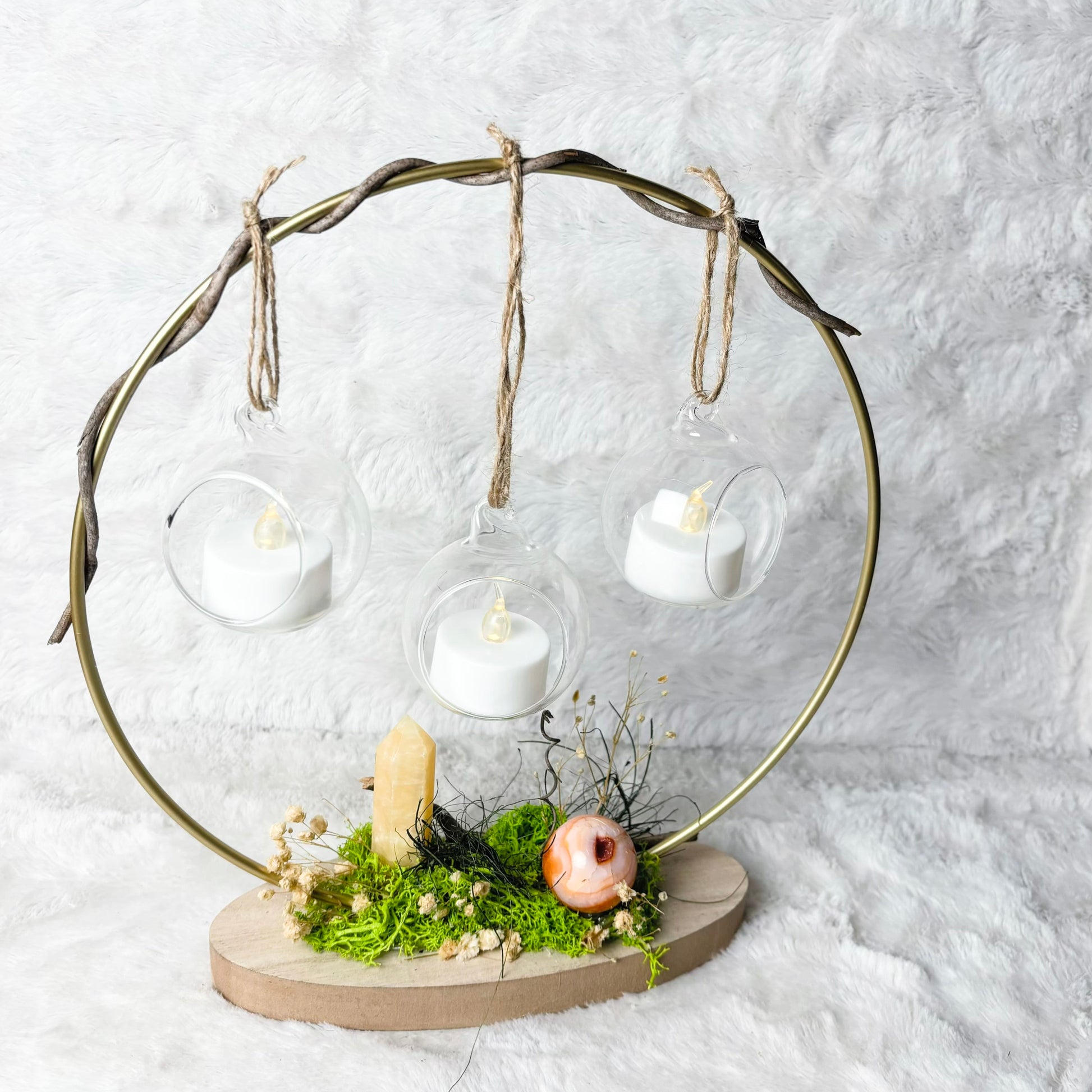 A decorative hanging candle display with three clear glass cups suspended from a twisted metal frame, set on a round wooden base with green moss, dried flowers, and small natural accents against a white textured background.