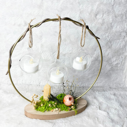 A decorative hanging candle display with three clear glass cups suspended from a twisted metal frame, set on a round wooden base with green moss, dried flowers, and small natural accents against a white textured background.