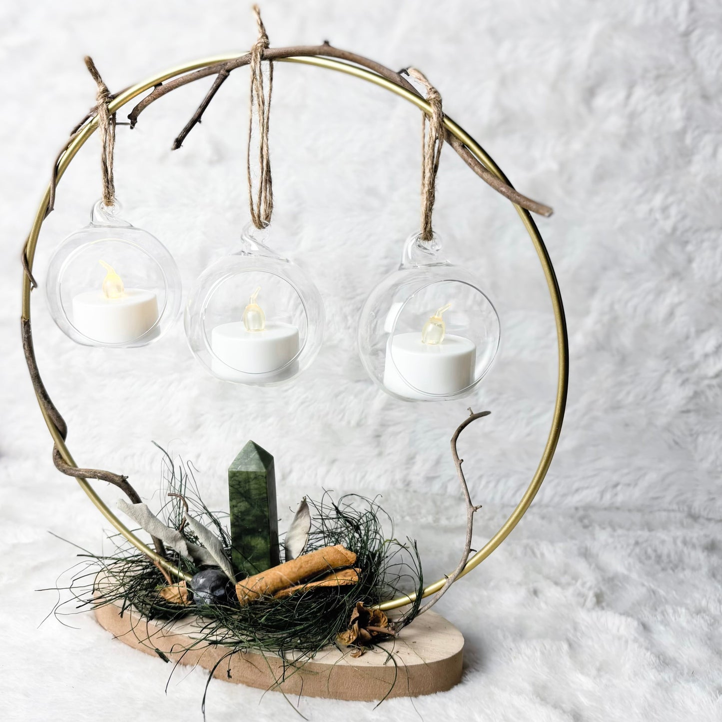 A decorative circular metal frame centerpiece with three hanging clear glass cups containing white tea light candles, mounted on a natural wood base with dried grass, a green crystal, and small natural accents against a soft white textured background.