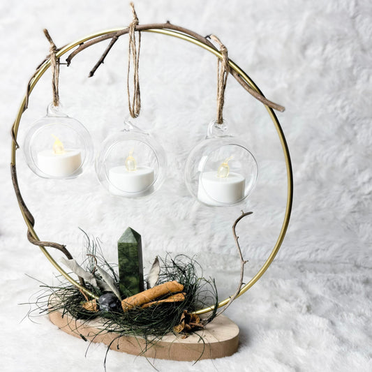 A decorative circular metal frame centerpiece with three hanging clear glass cups containing white tea light candles, mounted on a natural wood base with dried grass, a green crystal, and small natural accents against a soft white textured background.