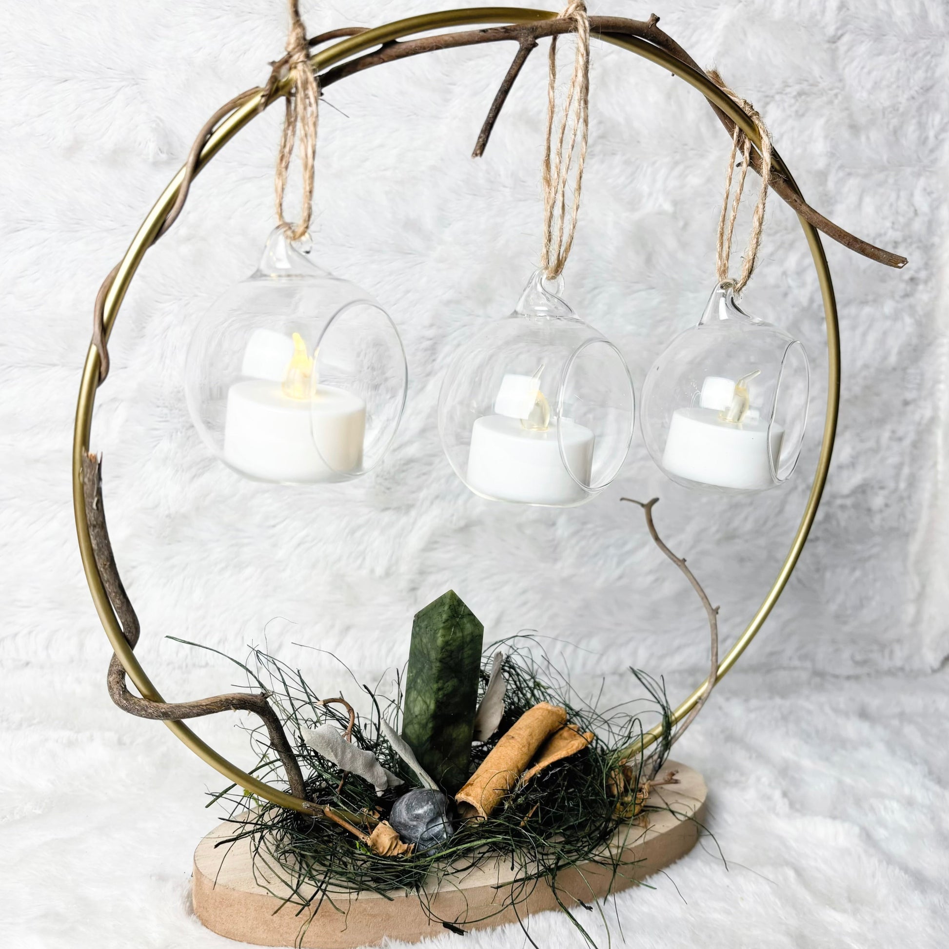 A decorative hanging arrangement made from a circular branch frame with three clear glass bell-shaped holders suspended by twine, each containing a white tea light candle. The base holds natural branches, moss, stones, and a green crystal on a round wood slice, set against a white textured backdrop.