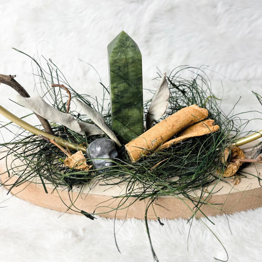 A natural dried floral arrangement centerpiece on a round light wood tray, featuring a green crystal tower, dried grasses, cinnamon sticks, a small gray stone, and branches, set against a white faux fur background.