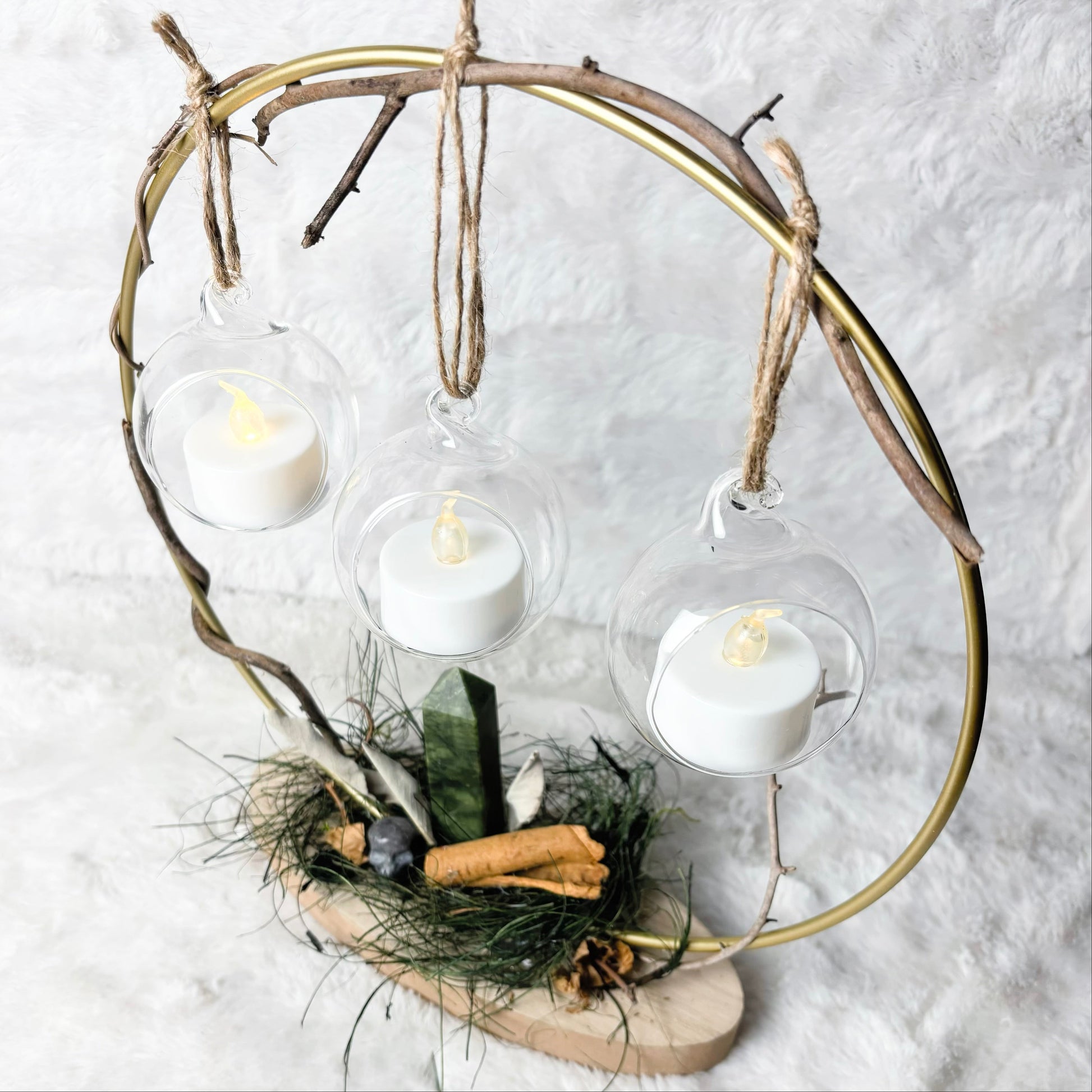 A rustic three-light hanging candle holder made from a circular branch frame and natural wood base, with three clear glass hanging bulbs containing white tea light candles, photographed on a white textured fabric background.