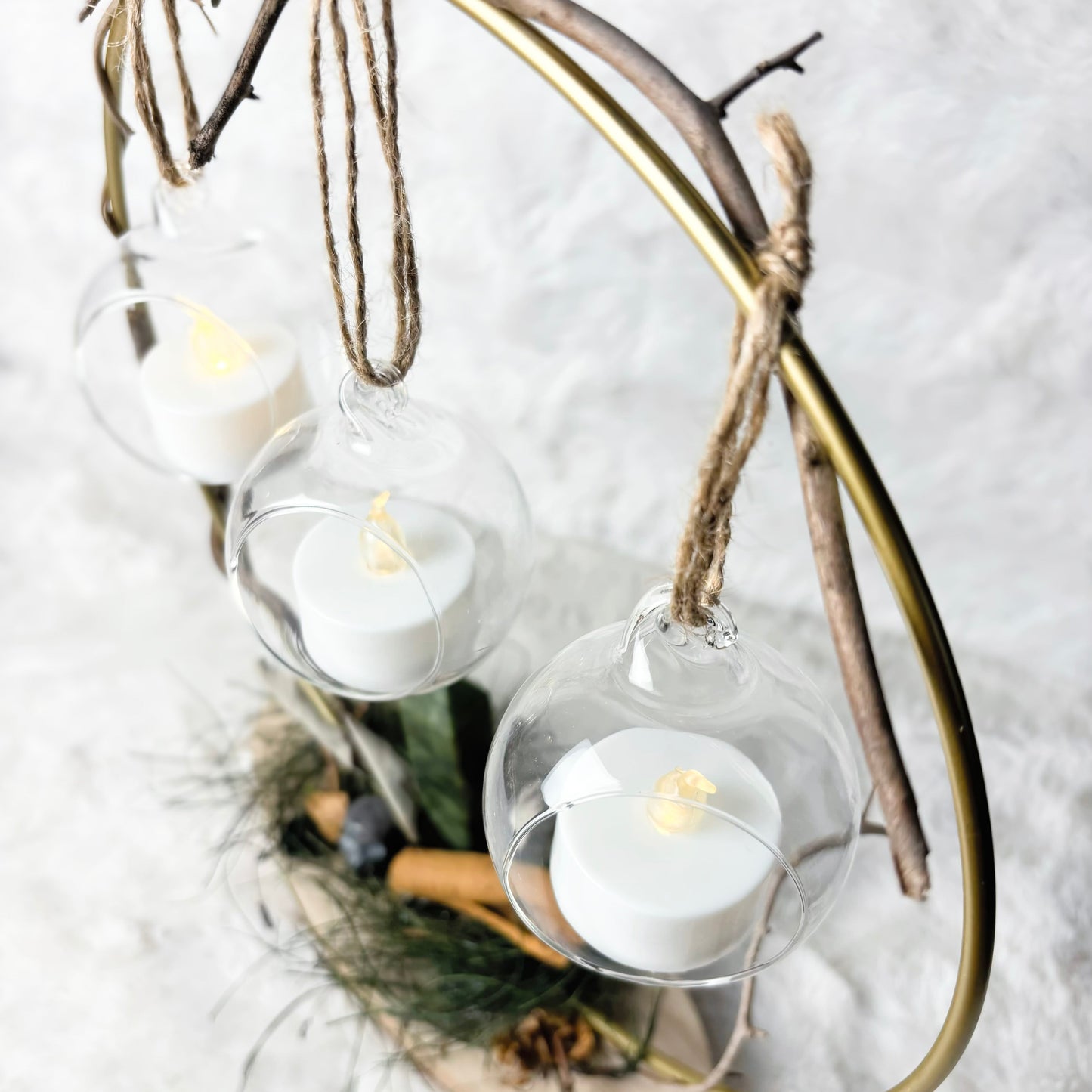 A rustic holiday hanging lantern with three clear glass cups suspended from a natural wood branch arch on a gold metal frame, each cup holding a white tea light candle with a yellow flame, set against a white textured background.