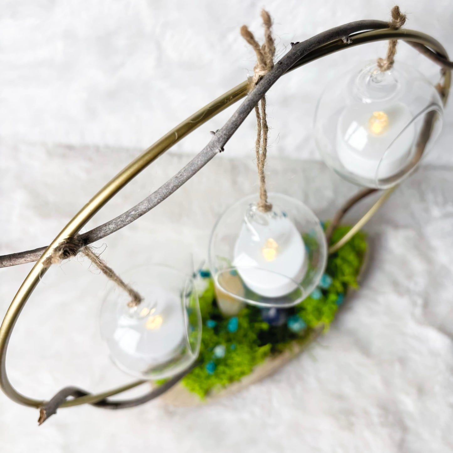 Three hanging glass teardrop lanterns with lit tea light candles, suspended from a rustic metal ring with twigs, resting on green moss and blue beads against a white textured background.