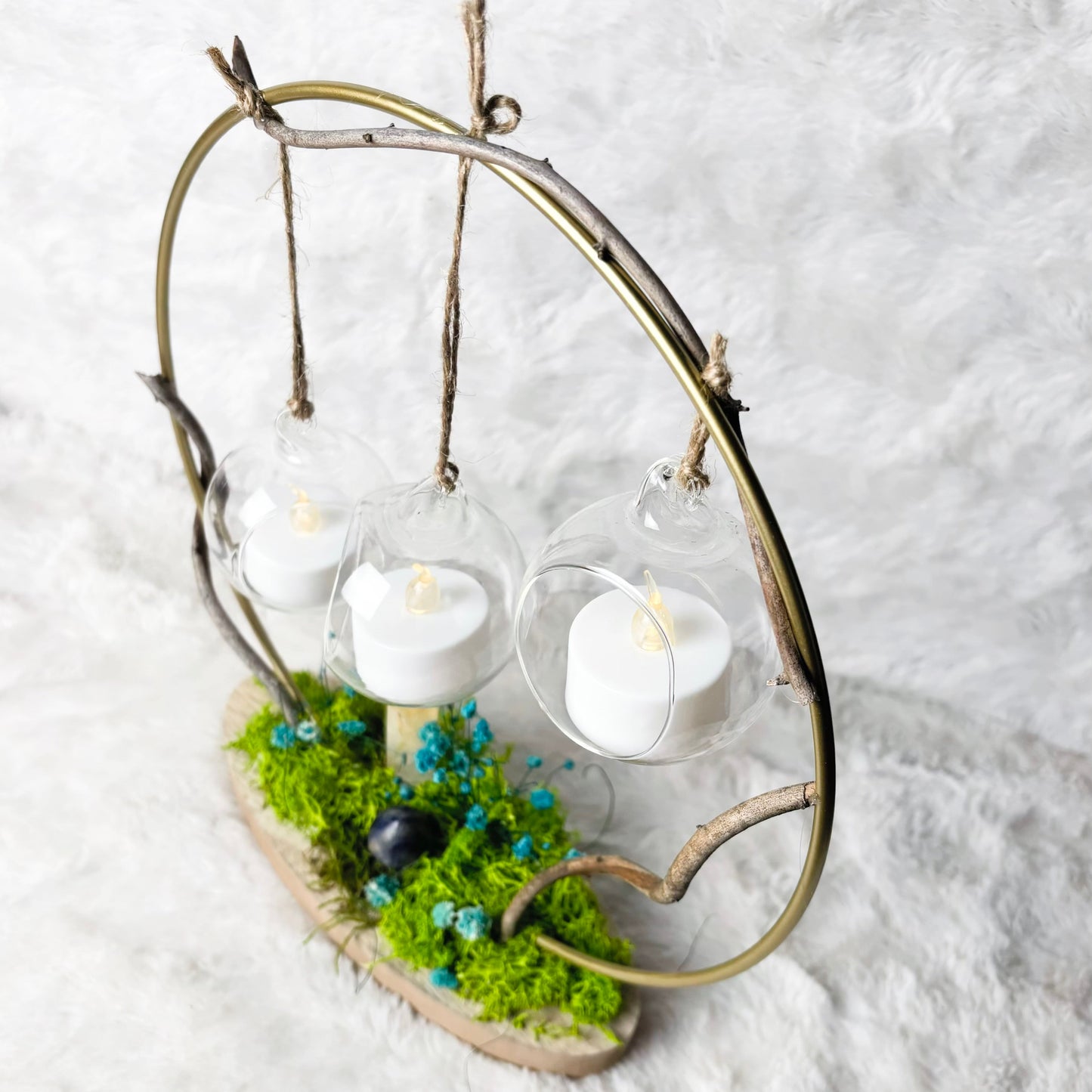 A decorative hanging candle holder made from a branch arch over a mossy base, holding three clear glass hanging spheres with white tea light candles, photographed on a white textured fabric background.