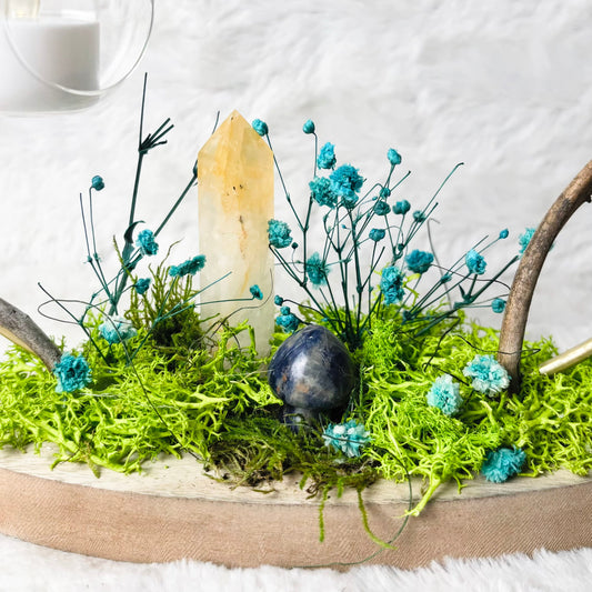 A decorative tabletop arrangement on a round wooden tray, featuring bright green moss, small blue flowers, a pale crystal, a dark blue stone, and two curved branches, styled on a white faux-fur surface with hanging glass candle holders in the background.