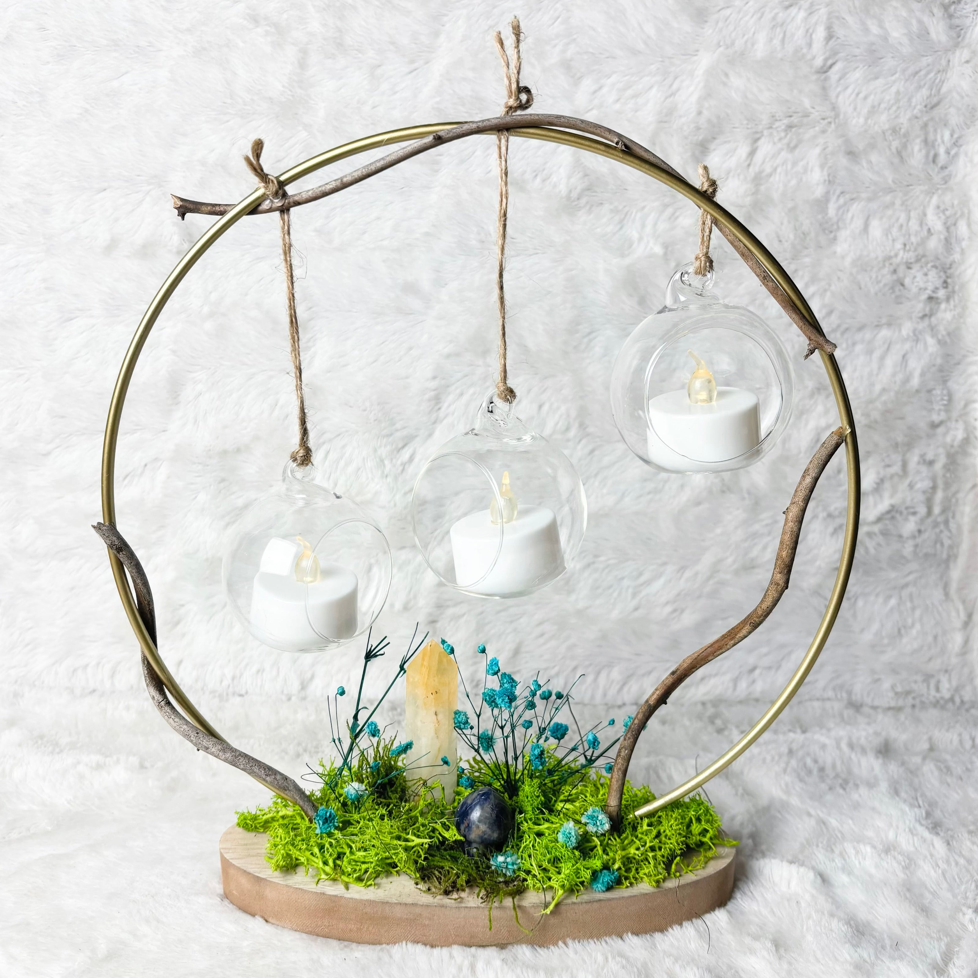 A decorative hanging candle holder made from a circular branch frame with three clear glass dome cups holding white tea light candles, mounted on a natural wood base with green moss and small blue wildflowers, photographed against a white textured fabric backdrop.