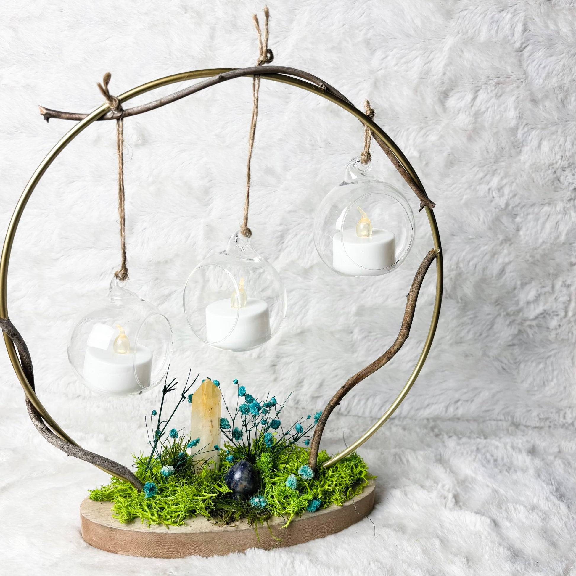 A decorative hanging candle display made from a circular branch frame with three clear glass globes holding white tea light candles, mounted on a natural wood base with green moss, small blue flowers, and a small bird figurine, set against a white faux-fur background.