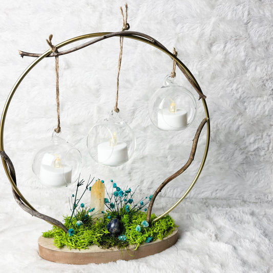 A decorative hanging candle display made from a circular branch frame with three clear glass globes holding white tea light candles, mounted on a natural wood base with green moss, small blue flowers, and a small bird figurine, set against a white faux-fur background.