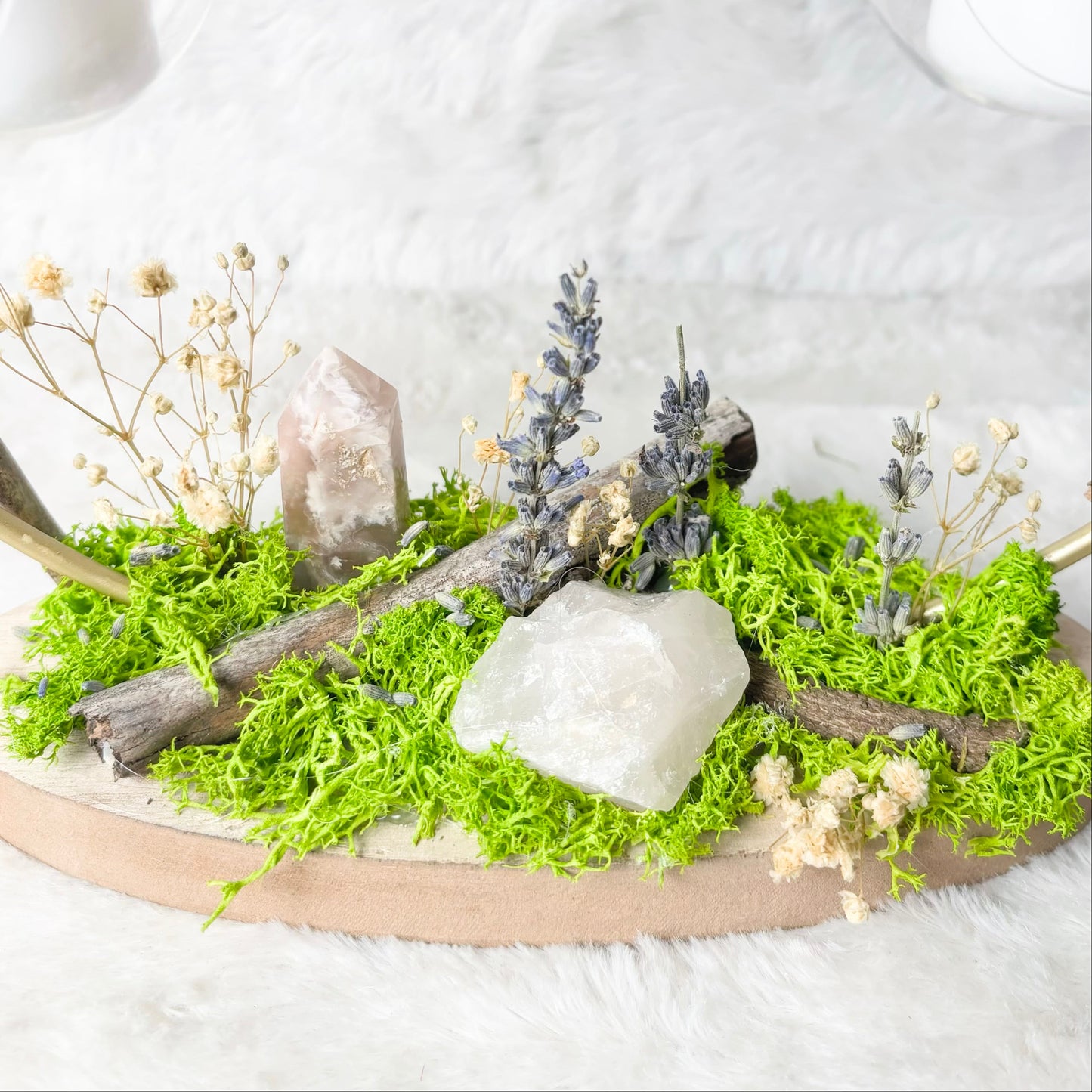 A decorative oval wooden tray filled with bright green moss, dried branches, small pale flowers, and natural stones, styled on a white faux-fur surface with blurred glass candle holders in the background.
