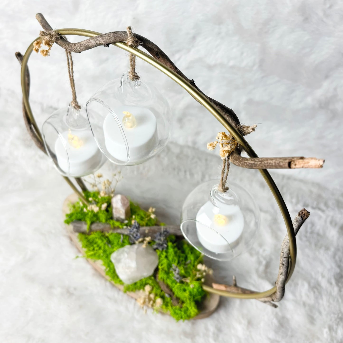 A decorative hanging light fixture made from natural branches and moss, holding three clear glass cups with white tea light candles, set against a soft white textured background.
