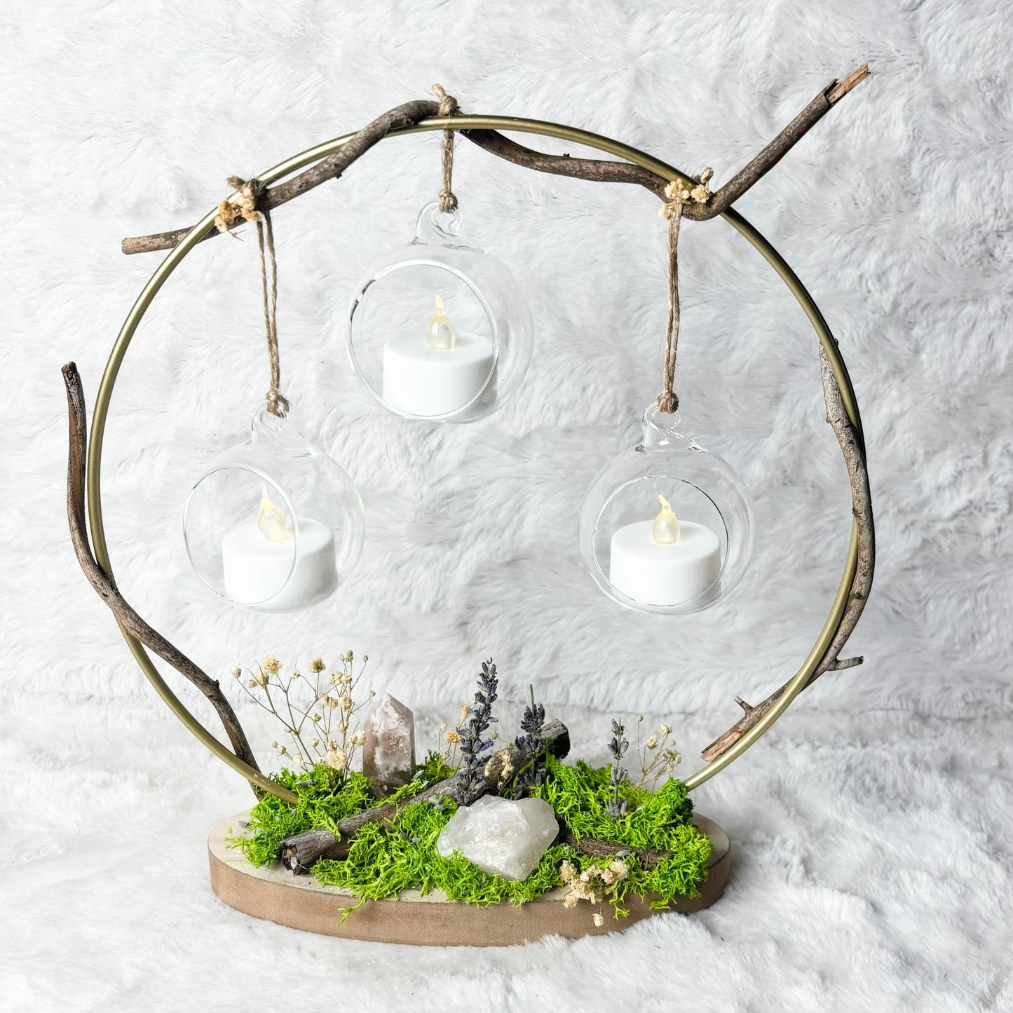 A rustic metal circular hanging frame with three clear glass cups holding white tea light candles, mounted on a natural wood base with green moss and small dried botanicals, set against a white textured fabric backdrop.