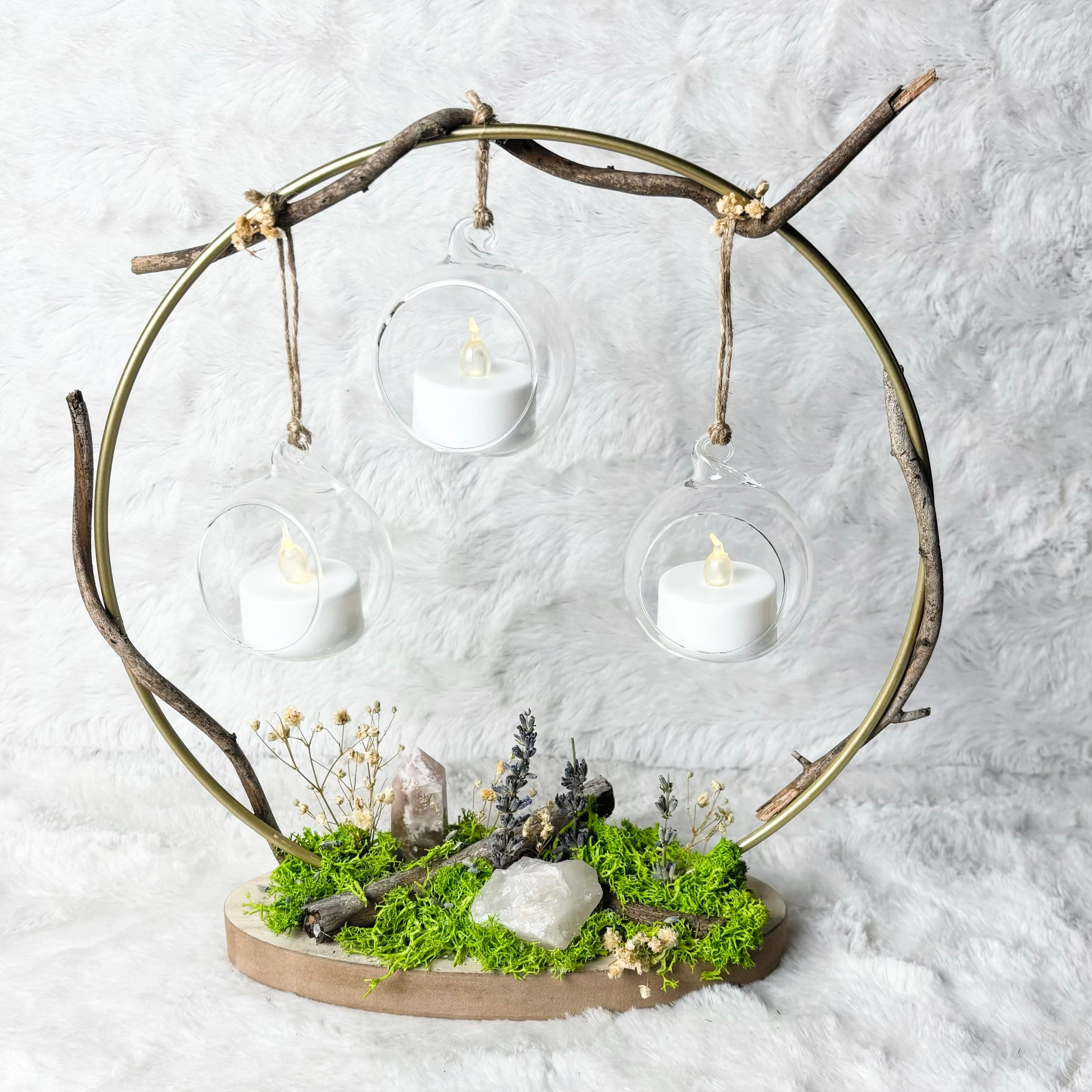 A rustic metal circular hanging frame with three clear glass cups holding white tea light candles, mounted on a natural wood base with green moss and small dried botanicals, set against a white textured fabric backdrop.