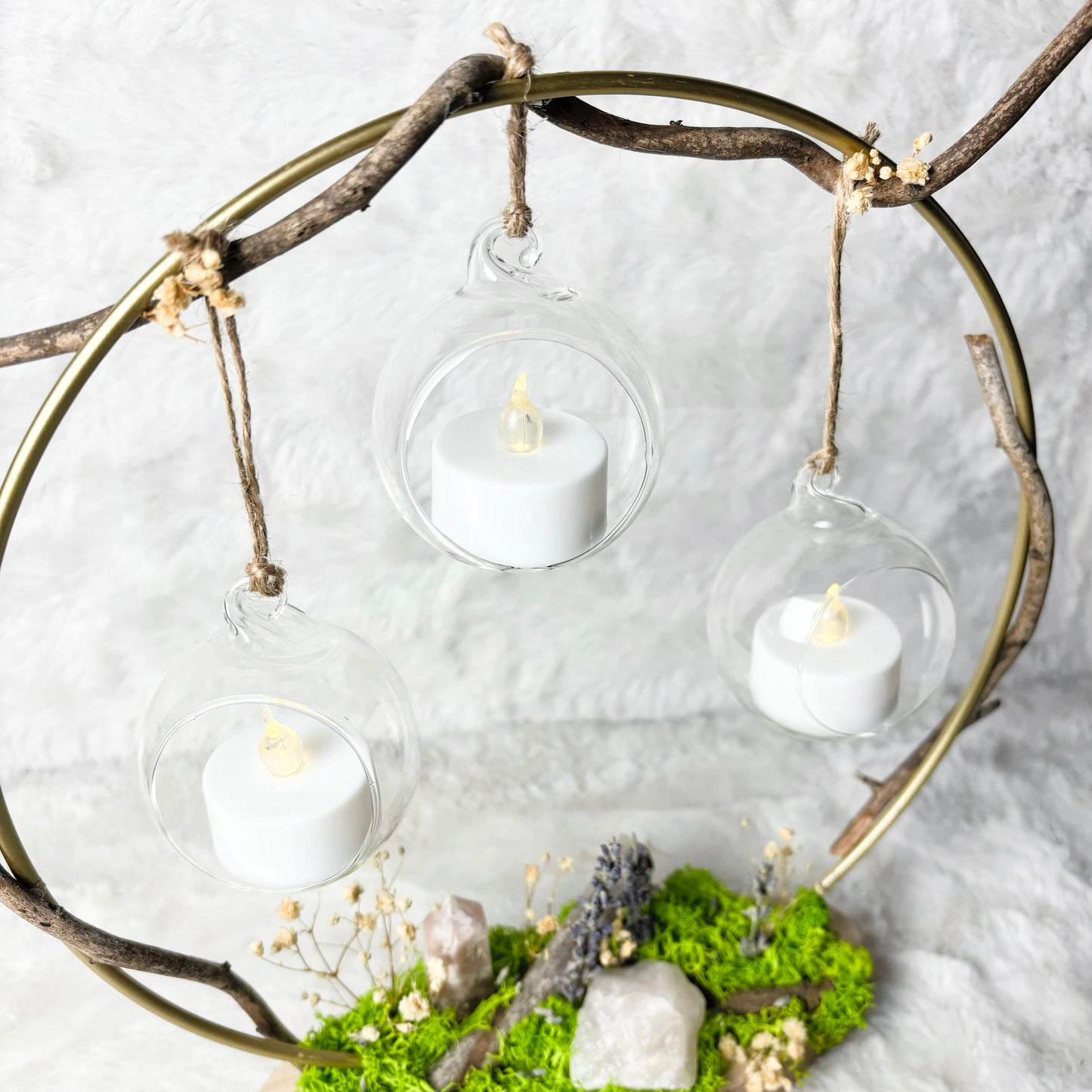 A decorative hanging arrangement with three clear glass orb lanterns containing white tea light candles, suspended from a rustic circular metal frame made of branches. The base features bright green moss, small stones, and dried floral accents on a light wood plaque, set against a soft white textured background.