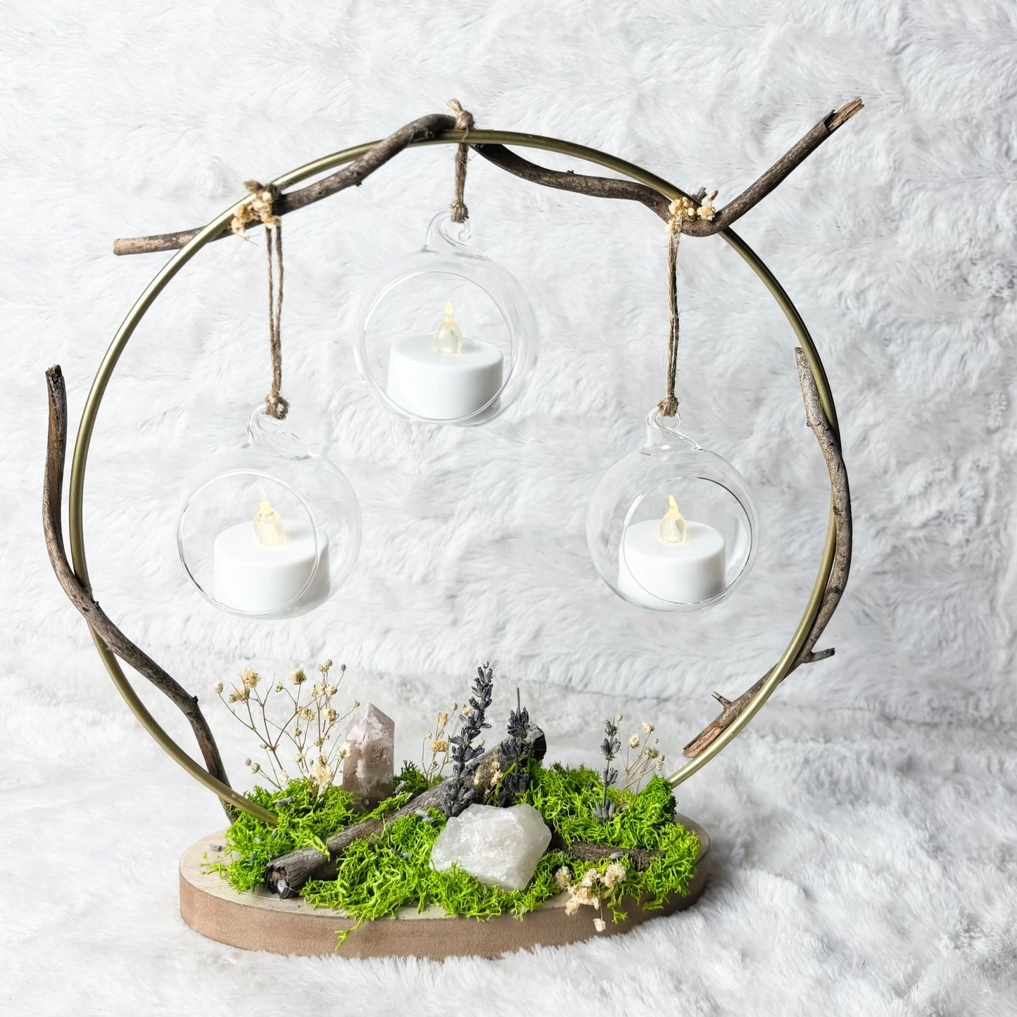 A decorative branch-shaped three-light holder with clear glass domes and white tea light candles, set on a mossy base with small stones and dried flowers, photographed against a white textured backdrop.