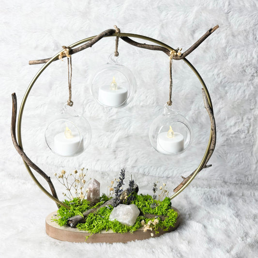 A decorative branch-shaped three-light holder with clear glass domes and white tea light candles, set on a mossy base with small stones and dried flowers, photographed against a white textured backdrop.