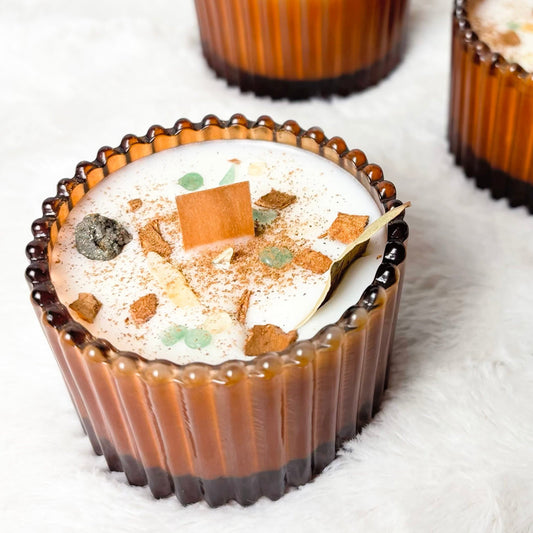 Three amber-brown ribbed glass cups filled with a creamy dessert topped with chocolate pieces, set on a white faux-fur surface.