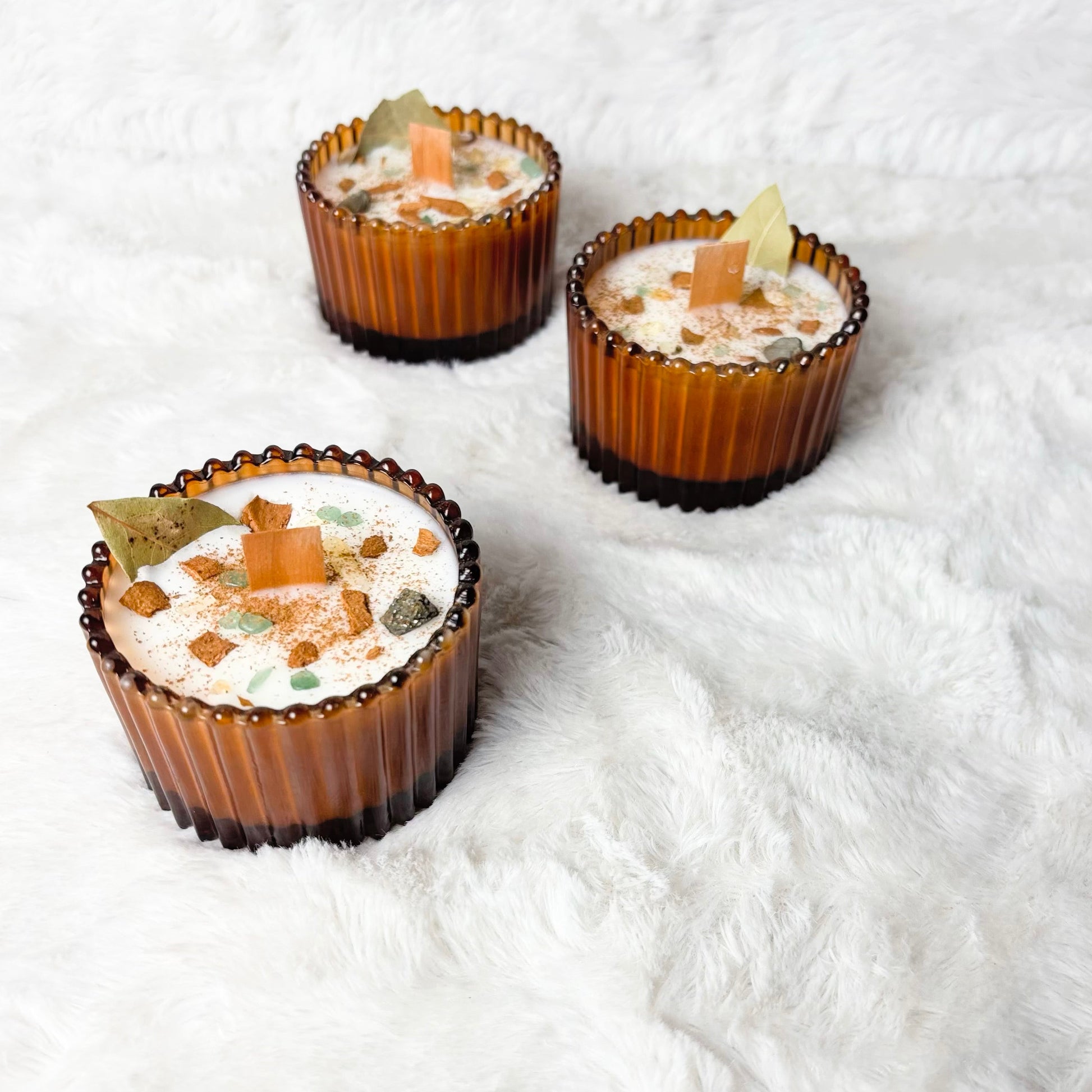 Three amber glass candle holders filled with white wax candles and topped with small decorative elements, arranged on a white faux fur surface.