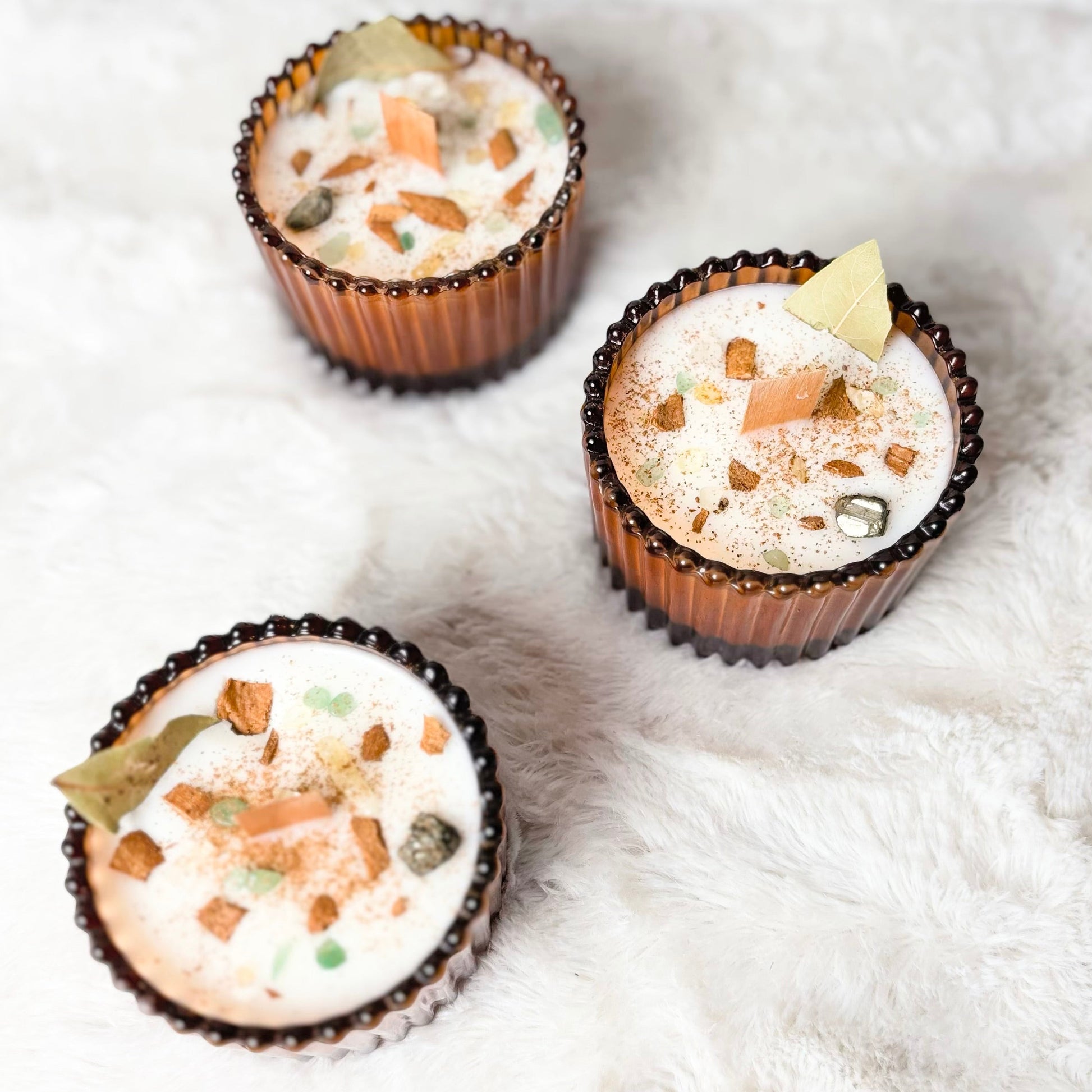 Three mini cupcakes in brown paper cups with white frosting, topped with nuts, cocoa powder, and a small vanilla piece, arranged on a white faux fur surface.