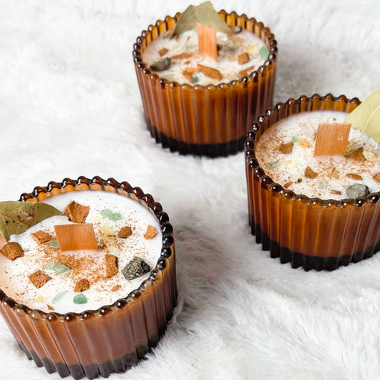 Three amber glass candle jars filled with white wax and topped with dried botanicals and small amber pieces, arranged on a white faux fur surface.