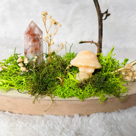 A decorative tabletop arrangement featuring bright green moss, a small beige mushroom figurine, a clear crystal point, dried flowers, a branch, and a glass candle holder with a white candle, set on a round wooden tray against a soft white background.