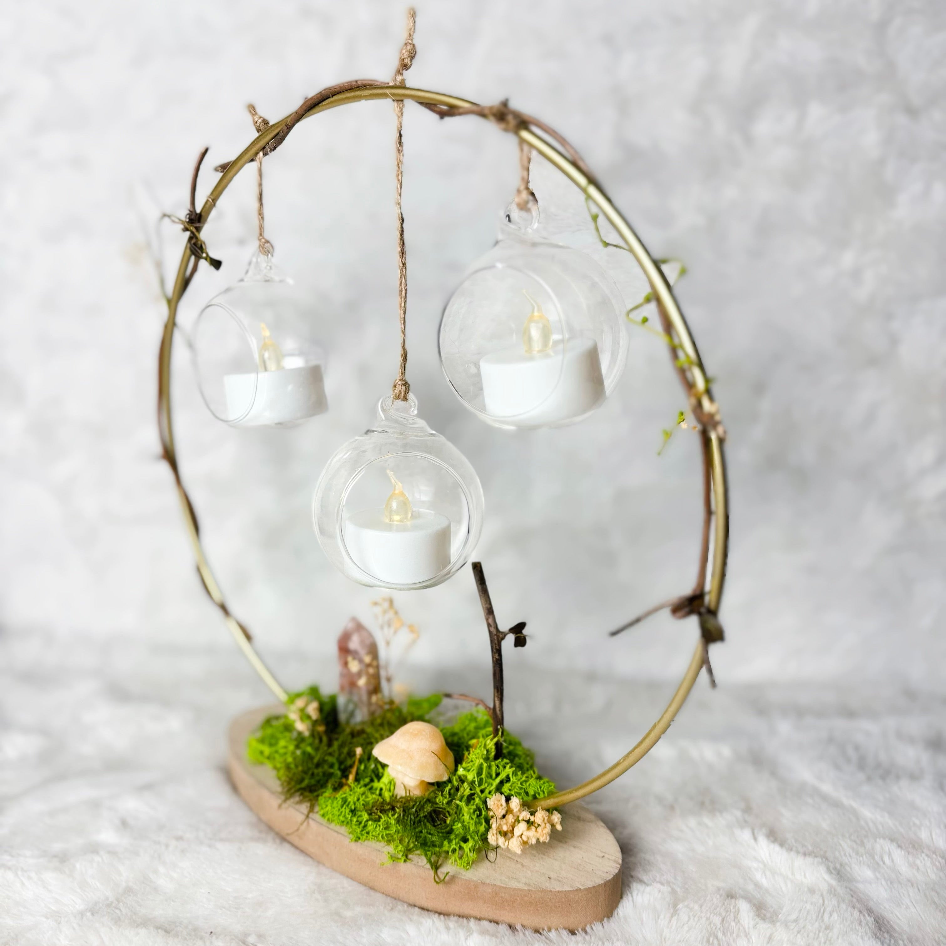 A decorative hanging lantern or fairy light display made from a circular branch frame, with three clear glass hanging bulbs containing white tea light candles, mounted on a wooden base with green moss and small dried flowers, set against a soft white textured background.