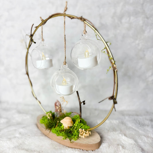 A decorative hanging lantern or fairy light display made from a circular branch frame, with three clear glass hanging bulbs containing white tea light candles, mounted on a wooden base with green moss and small dried flowers, set against a soft white textured background.