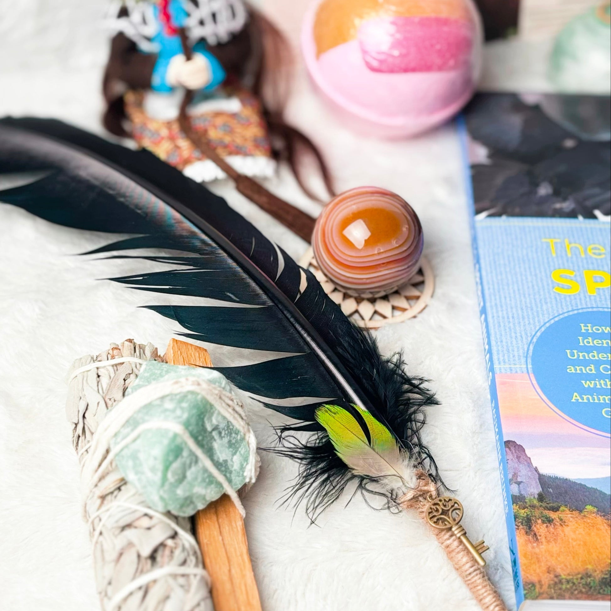 A styled flat-lay of spiritual and self-care items on a white textured surface, featuring a black feather, a smudge bundle with green and blue stones, a pink and orange bath bomb, a small owl figurine, and a book.