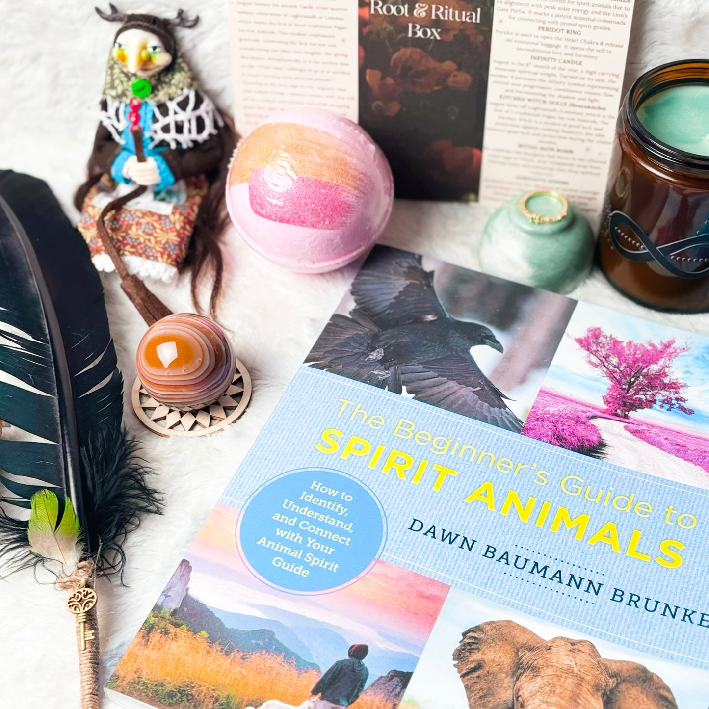 A styled flat-lay featuring the book 'The Beginner's Guide to Spirit Animals' by Dawn Baumann Brunke, surrounded by cozy decorative items including a feather quill, a pink bath bomb, a candle, and other books on a white faux fur surface.