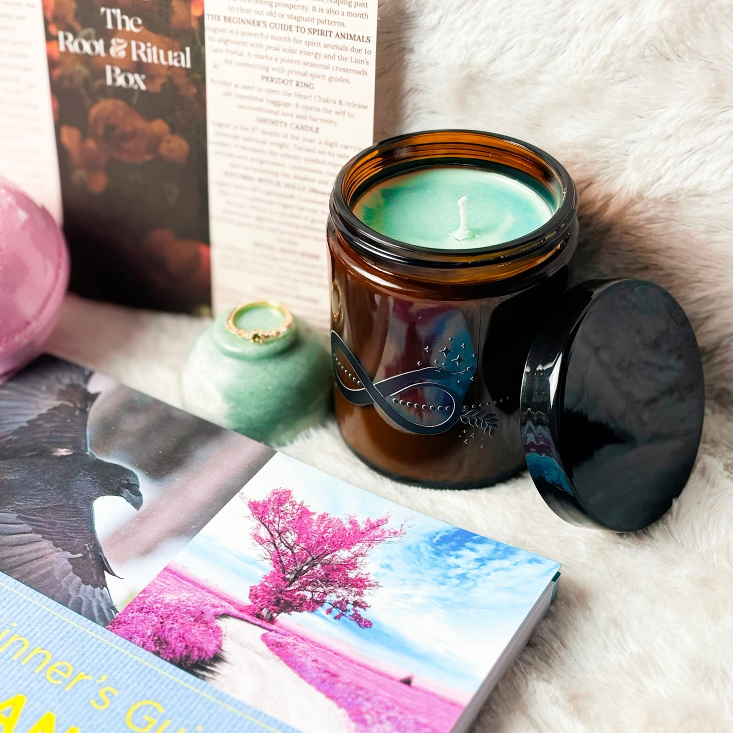 A styled flat-lay featuring a lit amber glass jar candle with a black lid resting beside it on a white faux fur surface, alongside a book titled 'The Root & Ritual Box' and another book partially visible at the bottom.