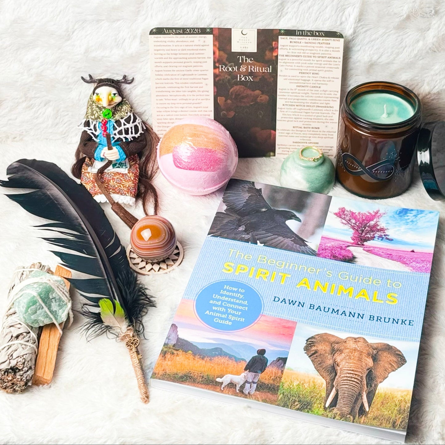 A styled flat-lay of a book titled 'Spirit Animals' by Dawn Baumann Brunke, surrounded by various spiritual and nature-themed props including a black feather, a smudge bundle, a crystal ball, a candle, and a small figurine, all arranged on a white textured blanket.