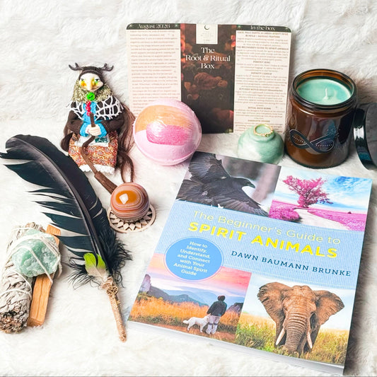 A styled flat-lay of a book titled 'Spirit Animals' by Dawn Baumann Brunke, surrounded by various spiritual and nature-themed props including a black feather, a smudge bundle, a crystal ball, a candle, and a small figurine, all arranged on a white textured blanket.