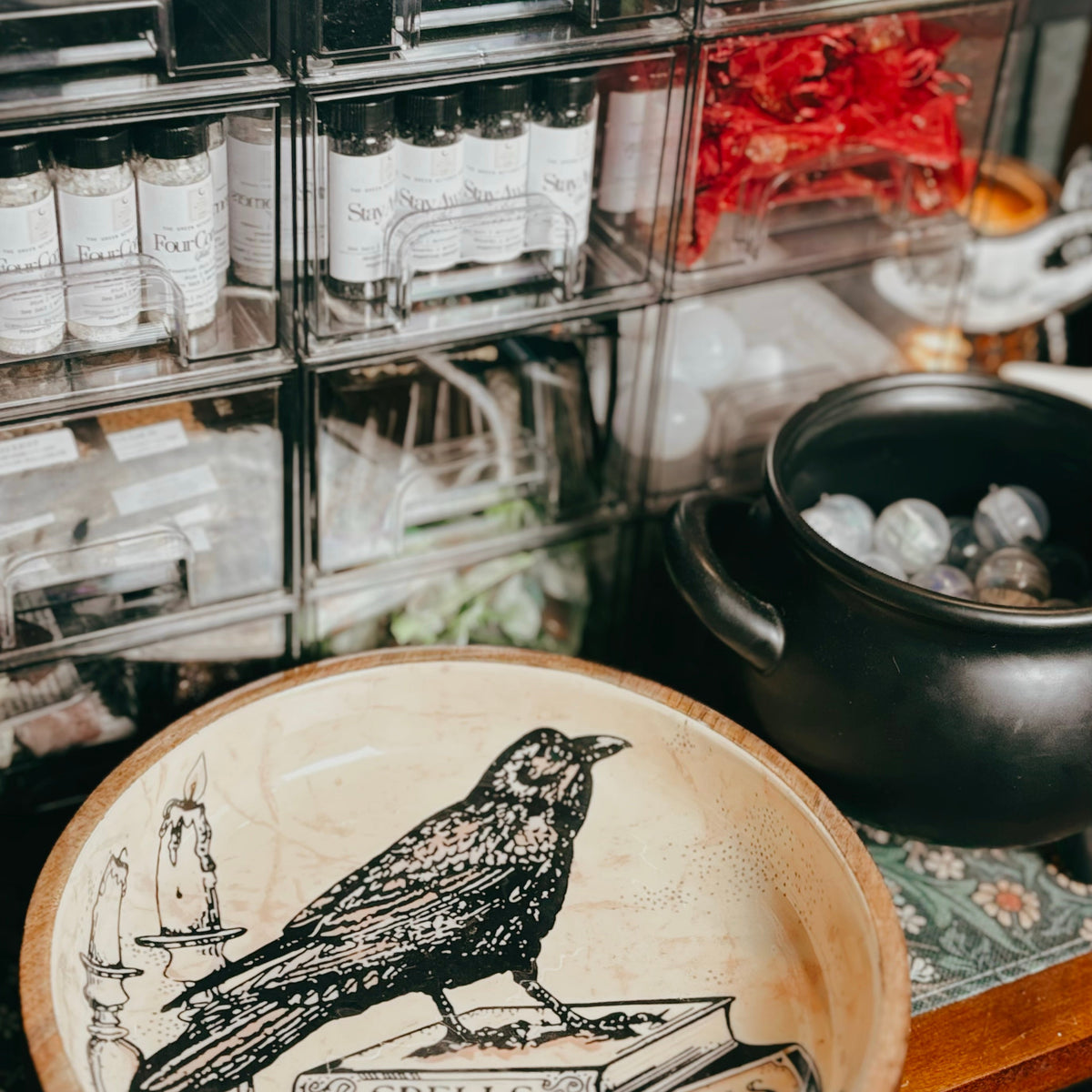 A close-up of a decorative ceramic bowl with a black raven illustration and the words 'SPELLS' and 'POTIONS' on a cream-colored surface, set in front of a shelf of small bottles and a black cauldron.