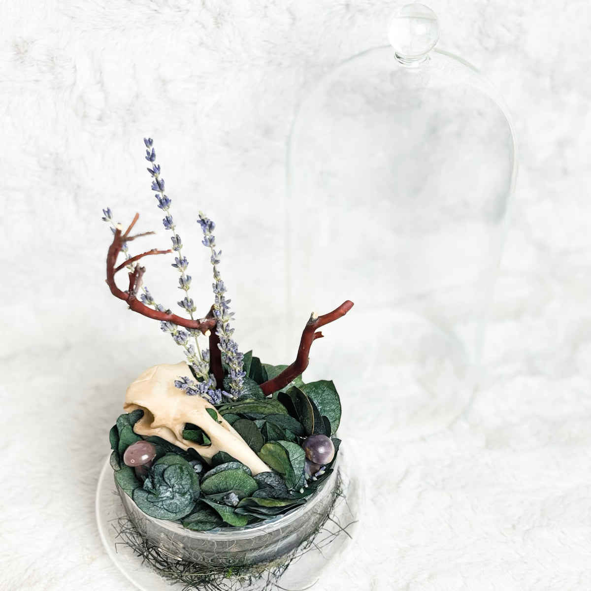 A decorative arrangement in a small metal bowl featuring green leaves, purple flowers, brown branches, and a cream animal skull, displayed on a white textured surface with a clear glass cloche behind it.